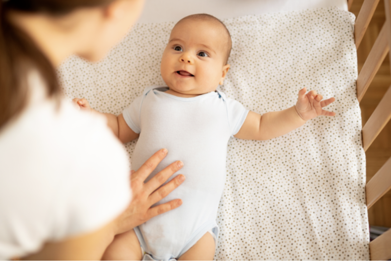 how to tell if baby is breathing properly. Parent leaning over side of crib and talking to her baby. Child looking at parent and smiling.